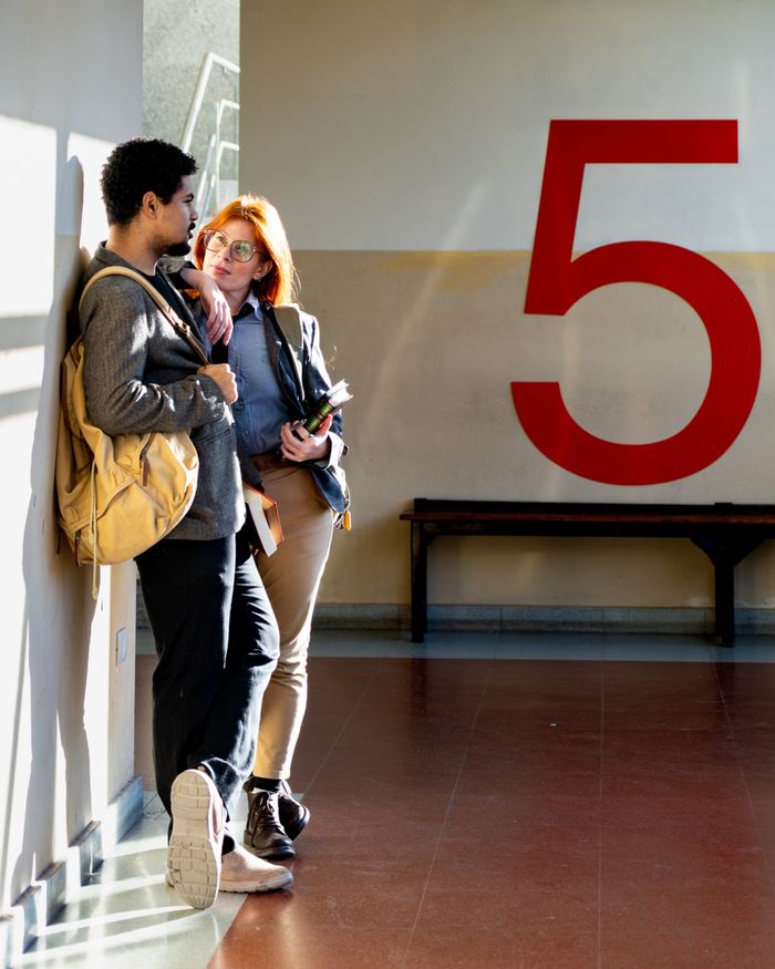 Two students discussing in a sunlit hallway in Buenos Aires, leaning against a wall marked '5'.